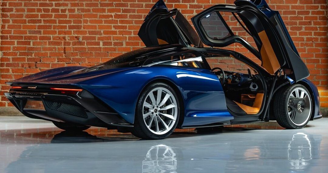 A sleek blue McLaren Speedtail with upward-opening doors is parked indoors on a shiny floor against a brick wall, showcasing its electrified longtail design.