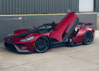 A red exotic car with an upward-opening driver door is parked on a concrete surface beside a gray industrial building, exemplifying the luxury cars offered by Velocity Motorcars.