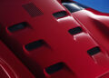 Close-up view of a red Ferrari car hood with multiple air vents, highlighting its aerodynamic design against a blue background.