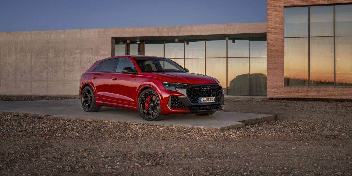 A red Audi RS Q8 Performance SUV is parked on a concrete platform in front of a modern building with large windows and brick accents, embodying powerful combustion at sunset.