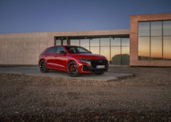 A red Audi RS Q8 Performance SUV is parked on a concrete platform in front of a modern building with large windows and brick accents, embodying powerful combustion at sunset.