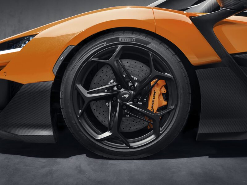 Close-up of a McLaren W1 sports car's front wheel, showcasing an orange brake caliper, sleek black alloy rim, and premium Pirelli tire against its glossy orange body. Check out the full specs and pricing for this remarkable design.