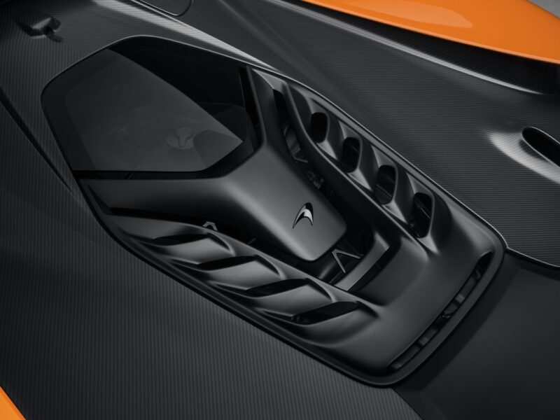 Close-up of an orange sports car's engine cover, featuring a sleek black design with aerodynamic vents resembling those in McLaren W1.