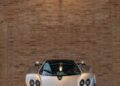 A sleek Pagani sports car is parked indoors against a brick wall, basking under the glow of a wall-mounted lamp.