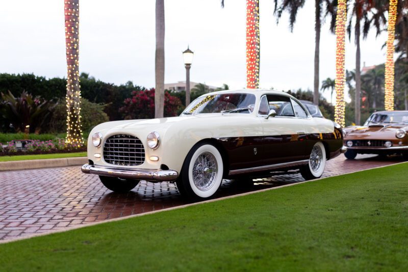 A vintage cream and brown car, reminiscent of Enzo Mattioli Ferrari's timeless designs, is parked on a brick road, surrounded by palm trees adorned with lights.