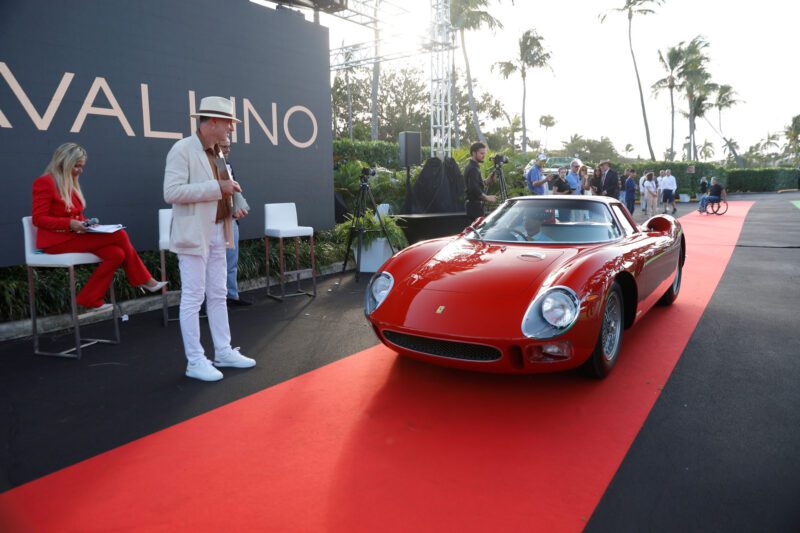 A red vintage Ferrari sports car gleams on the red carpet, captivating onlookers. Among the admirers stands a person in white attire, possibly someone from Cavallino Inc., adding an air of sophistication to this elegant scene.