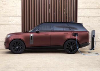 A maroon electric SUV from JLR is parked and plugged into a charging station against a beige wall, highlighting the shift towards hybrid sales in anticipation of FY25.