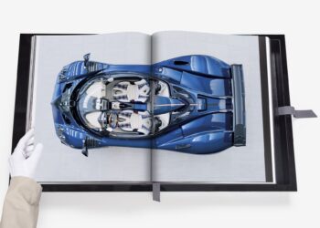 A gloved hand holds open an Assouline book, displaying an image of a blue sports car from a top-down view, showcasing one of its rare cars.
