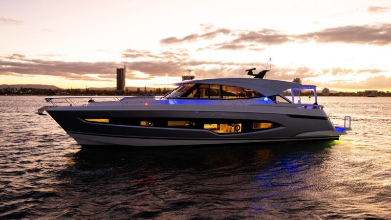 A luxury yacht glides on the water at sunset, silhouetted against a city skyline and a cloudy sky. Experience the allure of these magnificent vessels at the 2024 Fort Lauderdale International Boat Show.