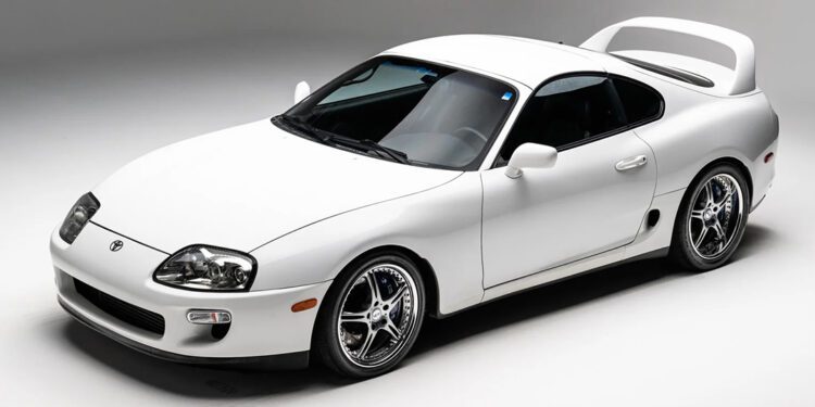 A white Toyota Supra, the crown jewel of JDM, boasts a sleek design and a large rear spoiler. Viewed from the front-left angle, it stands out against a plain background.