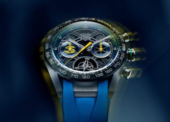 Close-up of a TAG Heuer Carrera Chronograph watch with a complex dial, visible mechanics, and a blue rubber strap on a blurred blue background.