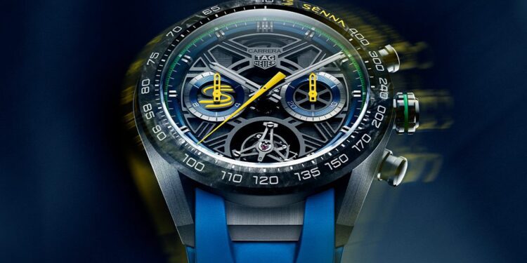 Close-up of a TAG Heuer Carrera Chronograph watch with a complex dial, visible mechanics, and a blue rubber strap on a blurred blue background.