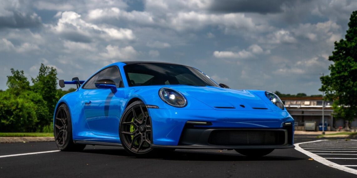 A bright blue exotic car from Velocity Motorcars is parked on an asphalt road, framed by a cloudy sky and lush greenery in the background.