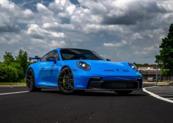 A bright blue exotic car from Velocity Motorcars is parked on an asphalt road, framed by a cloudy sky and lush greenery in the background.