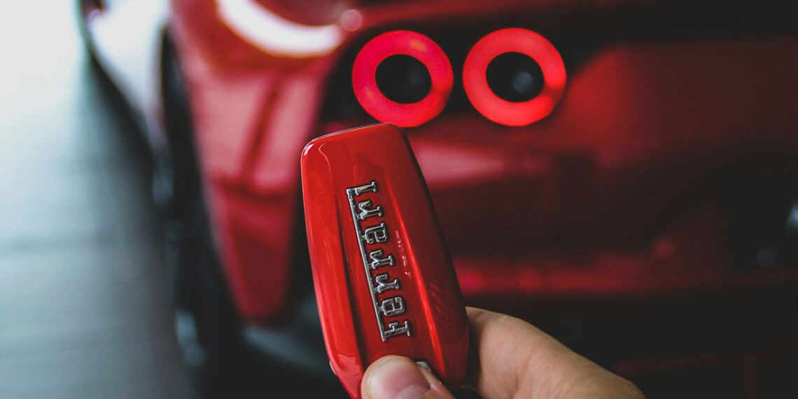 A close-up of a hand holding a red Ferrari key fob, with the blurred rear section of this exotic car showcasing its illuminated tail lights in the background.