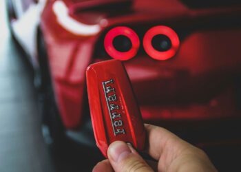 A close-up of a hand holding a red Ferrari key fob, with the blurred rear section of this exotic car showcasing its illuminated tail lights in the background.