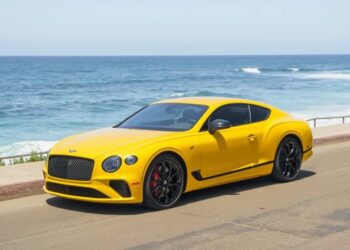A yellow sports coupe, reminiscent of the best Bentleys, is parked on a coastal road with the ocean and waves visible in the background.