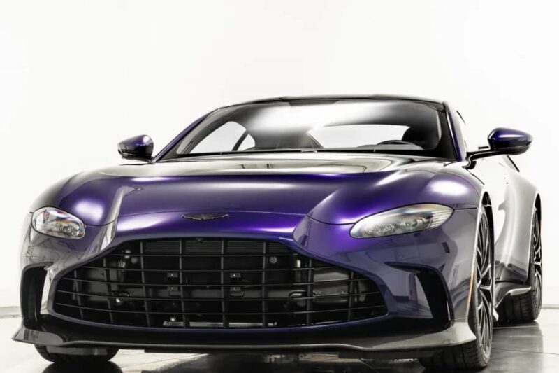 A front view of a 2023 Aston Martin V12 Vantage sports car with a low stance, wide grille, and sleek headlights, parked indoors against a plain white background.