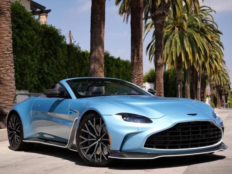A sleek 2023 Aston Martin V12 Vantage convertible in light blue is parked on a sunny street lined with palm trees, showcasing the perfect setting to buy Aston Martin and enjoy luxury under the sun.