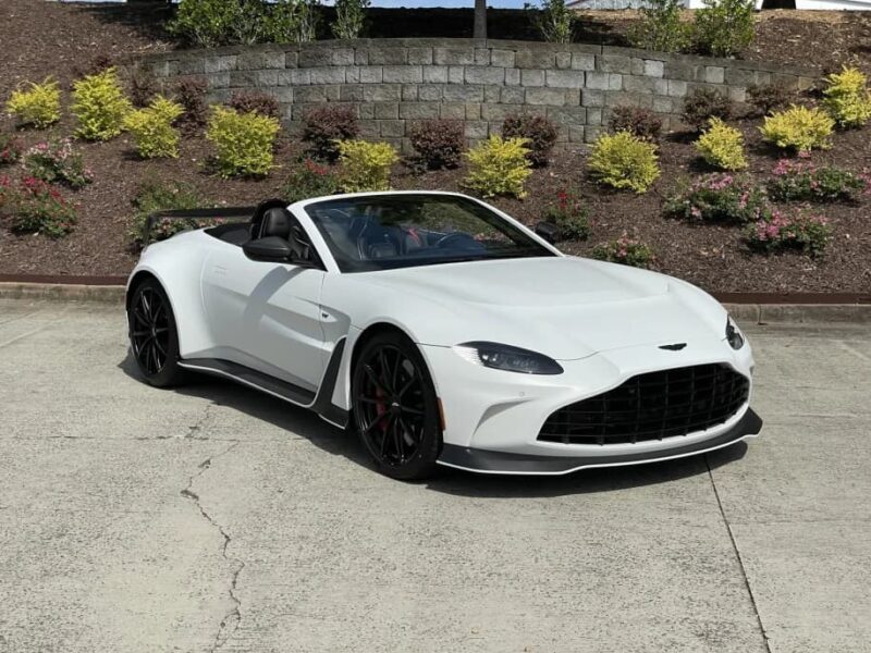A 2023 Aston Martin V12 Vantage convertible sports car is parked on a concrete surface in front of a landscaped area with shrubs and a stone retaining wall.
