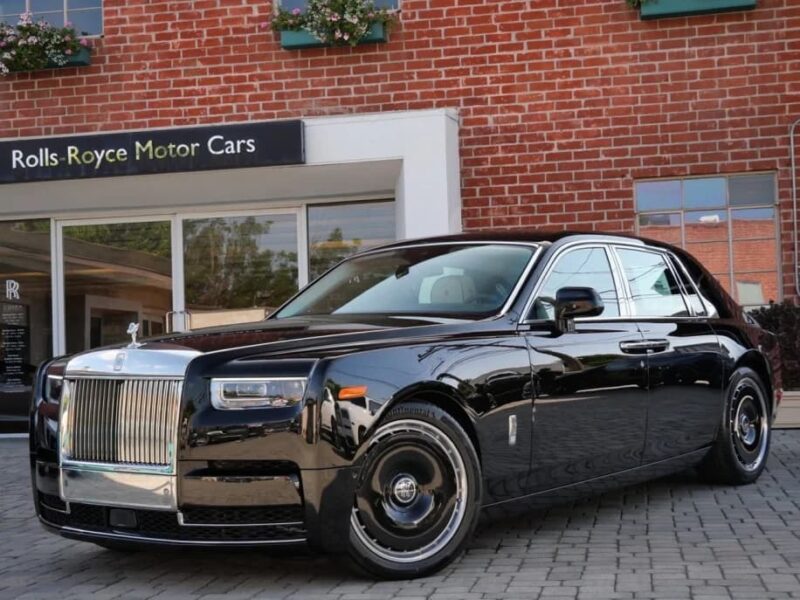 A black Rolls-Royce Phantom, the quintessential luxury sedan, is parked on a brick driveway in front of a Rolls-Royce Motor Cars dealership.
