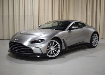 A silver 2023 Aston Martin Vantage coupe is parked indoors on a light-colored floor with beige curtains in the background.