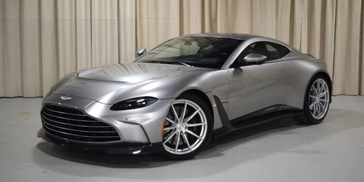 A silver 2023 Aston Martin Vantage coupe is parked indoors on a light-colored floor with beige curtains in the background.