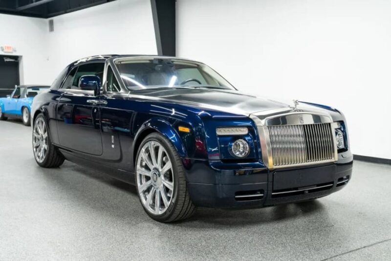 A quintessential dark blue Rolls-Royce Phantom coupe with chrome accents and large alloy wheels is parked indoors on a light gray floor.
