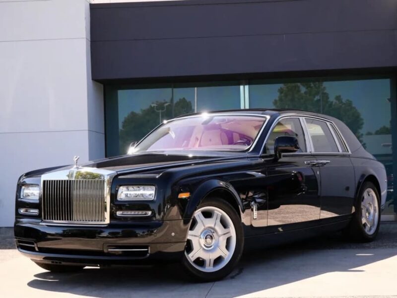 A black Rolls-Royce Phantom luxury sedan is parked in front of a modern building with large windows and a white-and-gray facade.