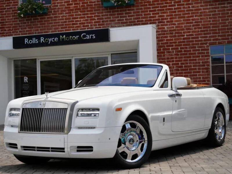 A white Rolls-Royce Phantom convertible, the epitome of a luxury car, is parked in front of a Rolls-Royce Motor Cars dealership with a classic brick exterior.