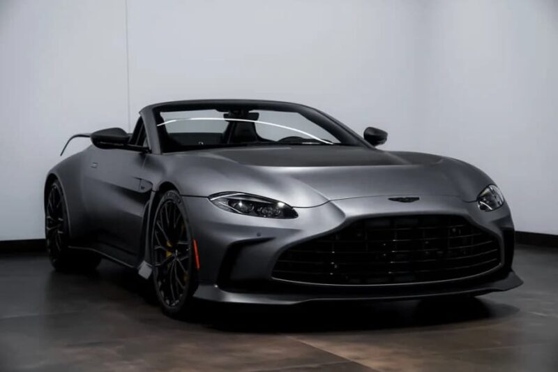 A matte black 2023 Aston Martin V12 Vantage convertible with the top down is parked indoors against a white wall.
