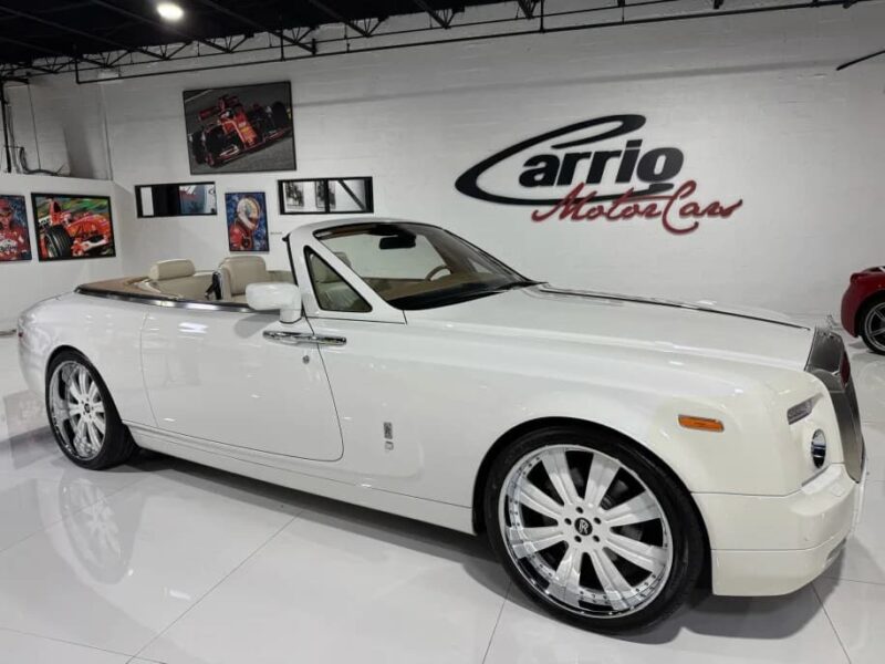 A quintessential white Rolls-Royce Phantom convertible with chrome wheels is parked indoors in a showroom with automotive-themed decor and gleaming white floors.