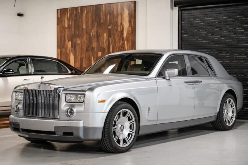 A quintessential Rolls-Royce Phantom luxury sedan in silver is parked indoors on a concrete floor, with another car partially visible in the background.