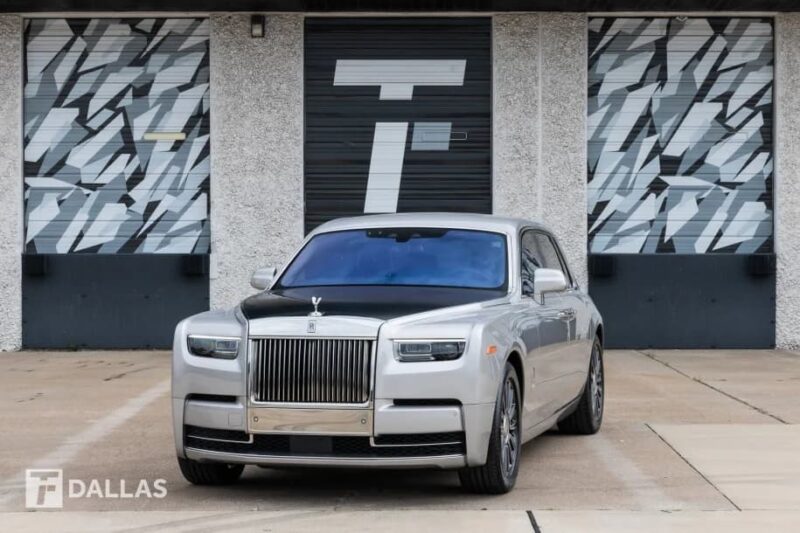 A quintessential silver Rolls-Royce Phantom luxury sedan is parked in front of a building with patterned garage doors and "T DALLAS" branding on the wall.