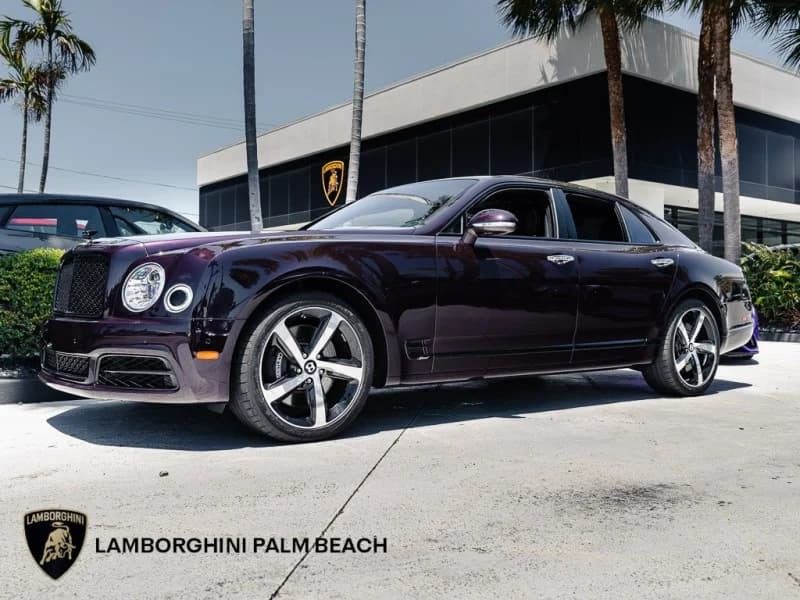 Dark purple sedan, one of the best bentleys for sale, is parked outside the modern agent building with palm trees and the Lamborghini Palm Beach brand.