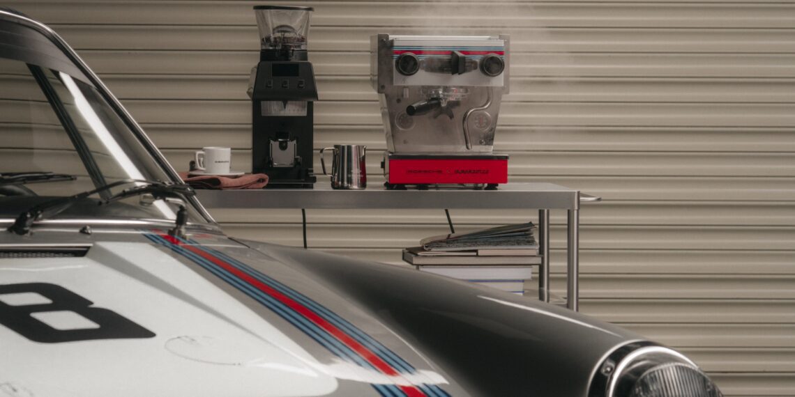 A sleek Porsche is parked in front of a table with a La Marzocco coffee machine emitting steam, a trusty coffee grinder, and books stacked cozily on one side.