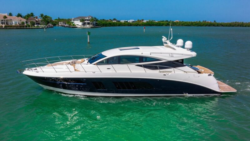 A white and black luxury yacht is sailing on clear turquoise water near a shoreline with houses and trees in the background, offering an unparalleled maritime experience.