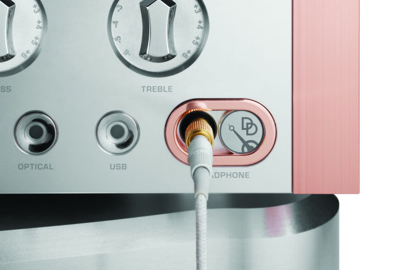 Close-up of a headphone jack plugged into a silver and pink Momentum C2 Preamplifier, featuring controls for bass, treble, optical, and USB—part of the finest audio system.