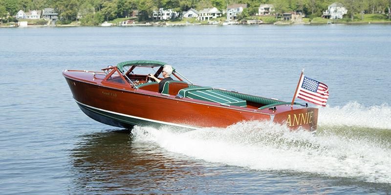 A wooden speedboat named "Annie" with a U.S. flag glides across a lake, offering a tranquil maritime experience, surrounded by houses and trees on the shoreline.