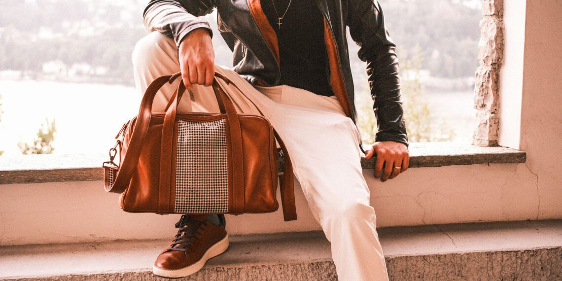 A person in a black Walter Leather Bespoke jacket and white pants sits on a ledge, holding a luxury brown leather duffle bag.