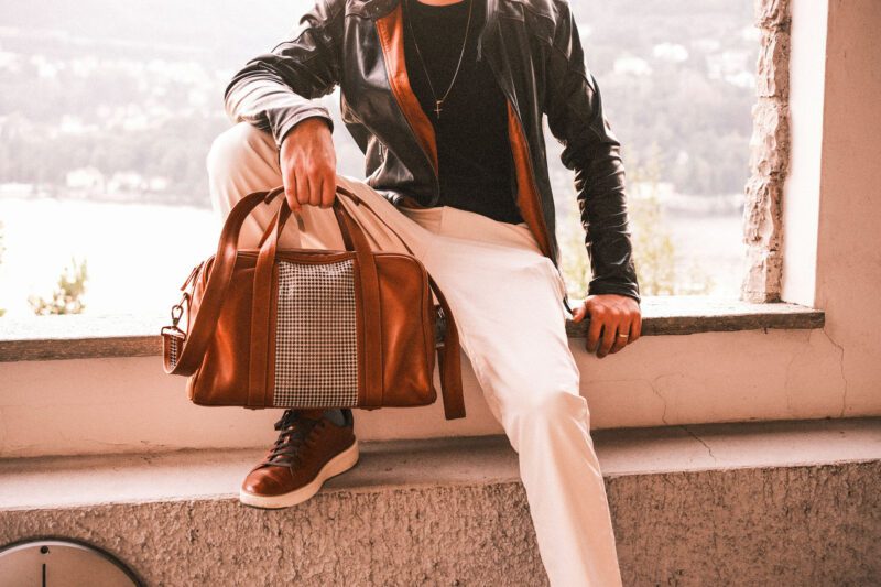 A person in a black Walter Leather Bespoke jacket and white pants sits on a ledge, holding a luxury brown leather duffle bag.
