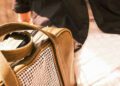 A person in a black coat sits next to a large olive green bag with a houndstooth-patterned pocket, reminiscent of custom leather luggage.