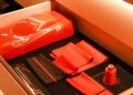 A drawer containing various red and black fabrics, a red thread spool, and a black folder labeled "Ferrari," all meticulously organized under warm lighting, reminiscent of jackets tailored to perfection.