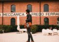 A person with a brown leather bag, reminiscent of Walter Leather Bespoke's craftsmanship, stands in front of a building labeled "Meccanica Alfredo Ferrari," while people are seated at white tables in the background.