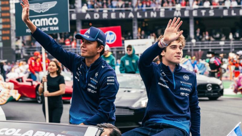 Two individuals in blue jackets wave from a vehicle during a public event, reminiscent of the vibrant atmosphere at an F1 starting grid. A crowd is visible in the background, capturing the excitement of events like the upcoming 2025 F1 Season.