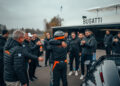 A driver in racing gear embraces another person in front of a Bugatti building, celebrating a world record. A group of people in matching jackets claps and watches, with the sleek W16 Mistral partially visible in the foreground.