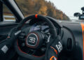 A person expertly handles a Bugatti W16 Mistral, racing at an astonishing 325 km/h on the track. The sleek dashboard and steering wheel are in sharp focus, with a vibrant tapestry of autumn trees rushing by in the background, hinting at a world record attempt.