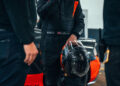 A race car driver in a black and orange suit holds a helmet by their side, standing near a Bugatti W16 Mistral in the garage, poised to chase the 282 MPH top speed world record.