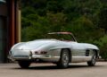 A classic 1957 Mercedes-Benz 300 SL convertible with a red interior is parked on the driveway, surrounded by greenery. This Rudge Roadster is for sale, offering a timeless blend of elegance and performance.