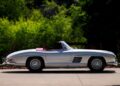 A vintage silver Mercedes-Benz 300 SL convertible car, showcasing a striking red interior, is parked gracefully on a driveway, enveloped by lush green foliage.